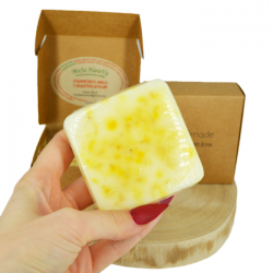 Roka drži svetlo rumeno glicerinsko milo z rumenimi cvetnimi lističi, v ozadju kartonasta škatlica